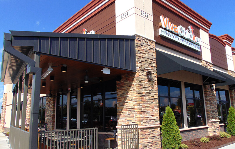 A restaurant with stone and tan walls, large windows, dark trim, and a covered outdoor patio. The sign reads